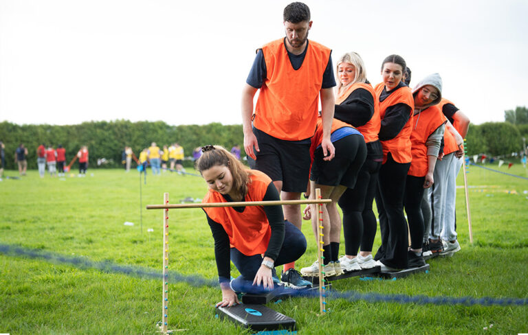 Team balancing on planks with woman ducking underneath limbo bar