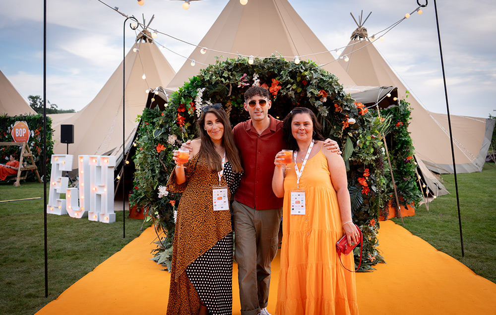 Three guests stood on an orange carpet posing in front of the floral tipi entrance